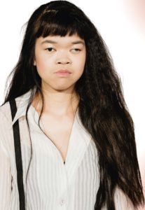 Die Schauspielerin und Musikerin Hieu Pham vom Theater RambaZamba. Sie ist Asiatin und hat lange, schwarze Haare mit Pony. Sie trägt eine weiße Bluse mit Streifen und Hosenträger und schaut ernst leicht an der Kamera vorbei.